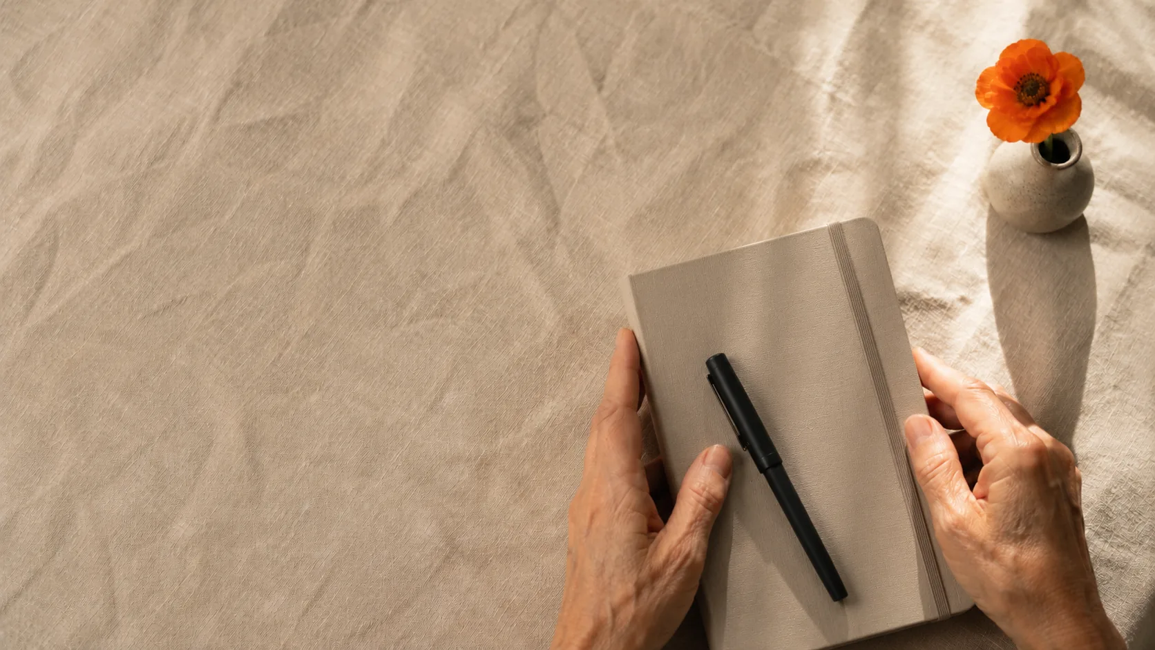 Hands holding a notebook and pen beside a small vase with an orange flower on warm linen