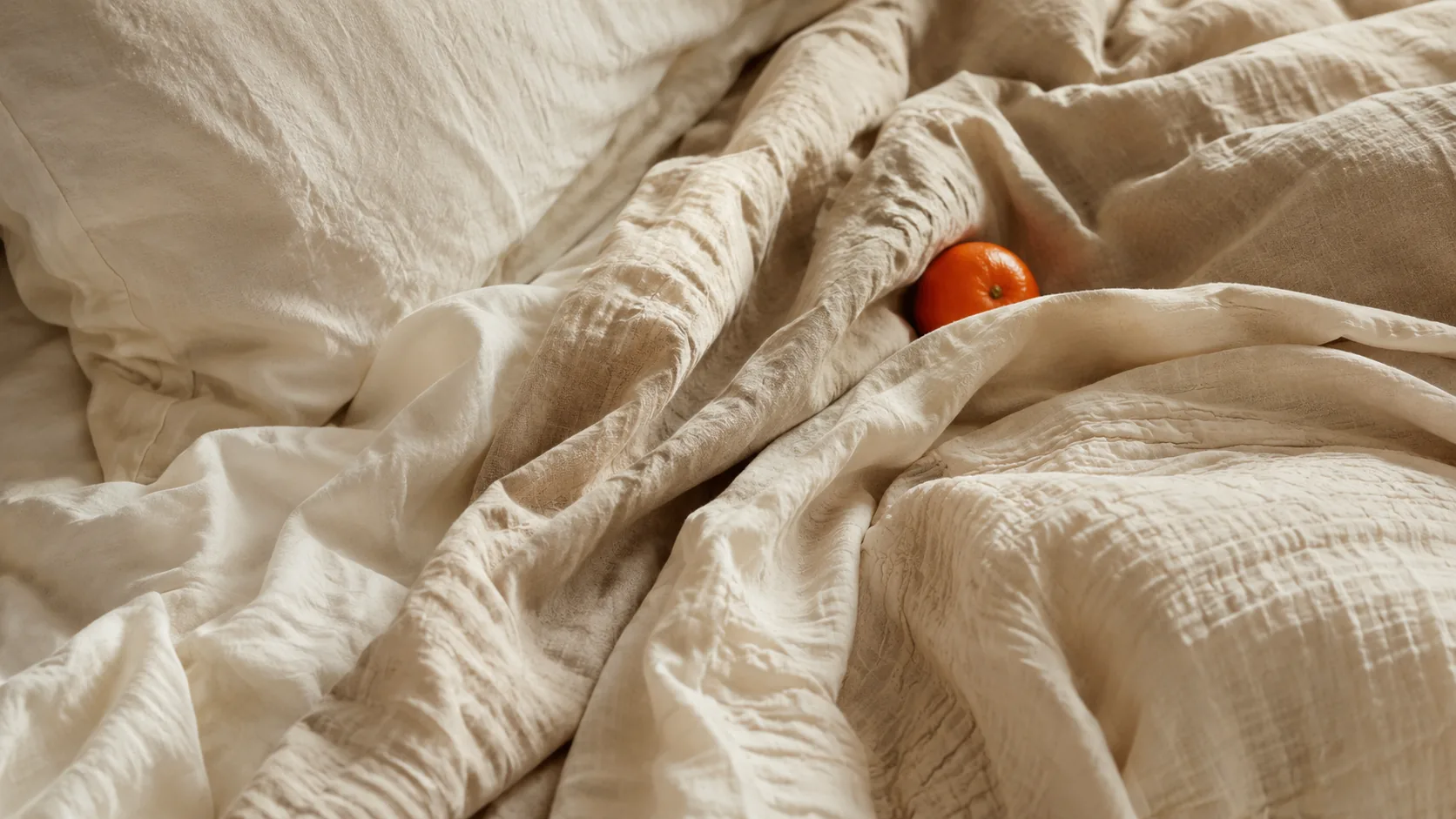 Sunlit linen sheets with a single clementine resting on the bed, in warm morning light.