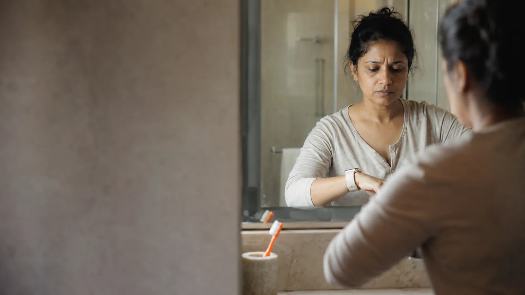 Woman frowns at her smartwatch in the bathroom mirror, anxious about her sleep data