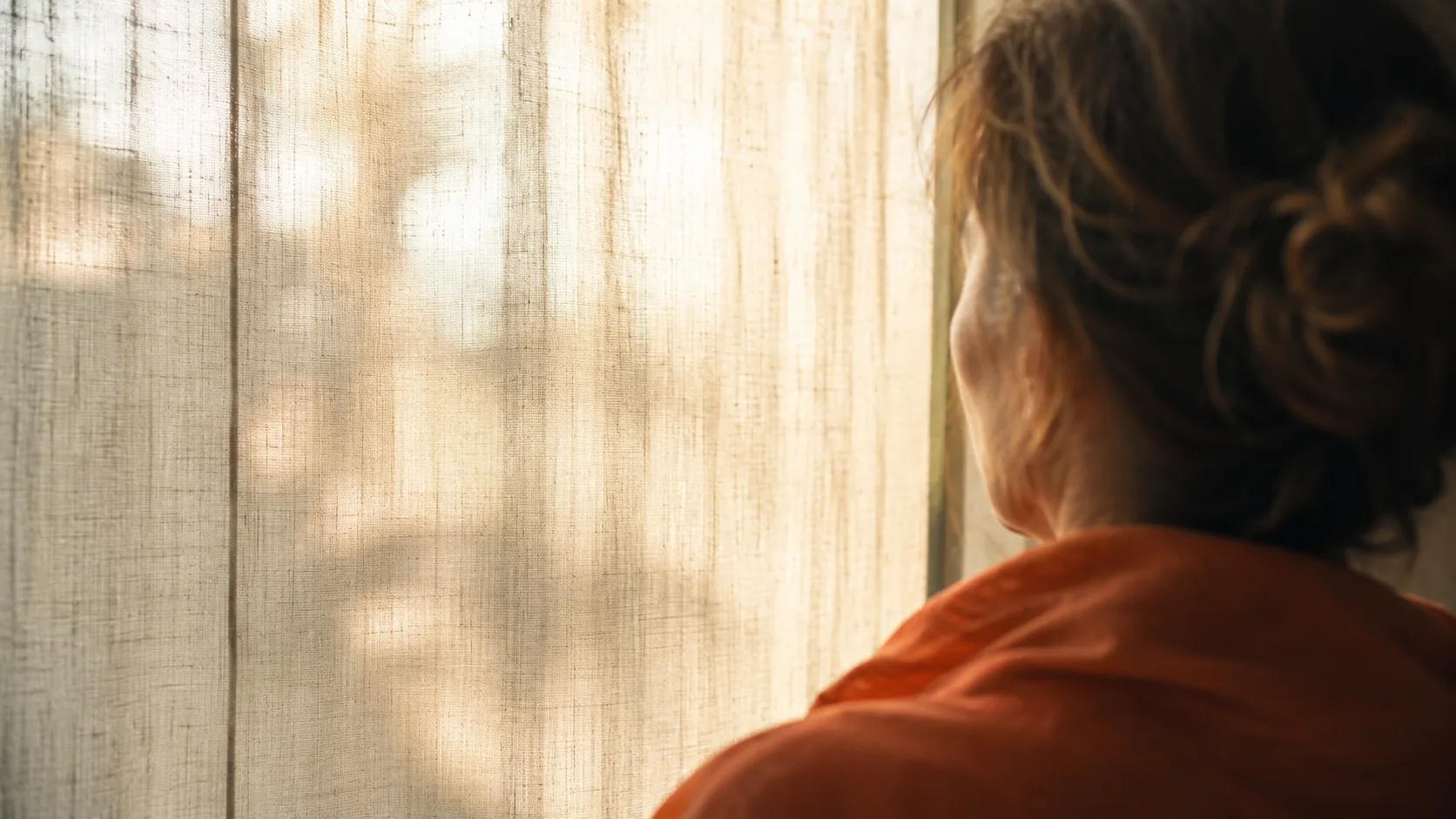 Woman wrapped in orange sits by a sunlit window, looking pensive as her mind races