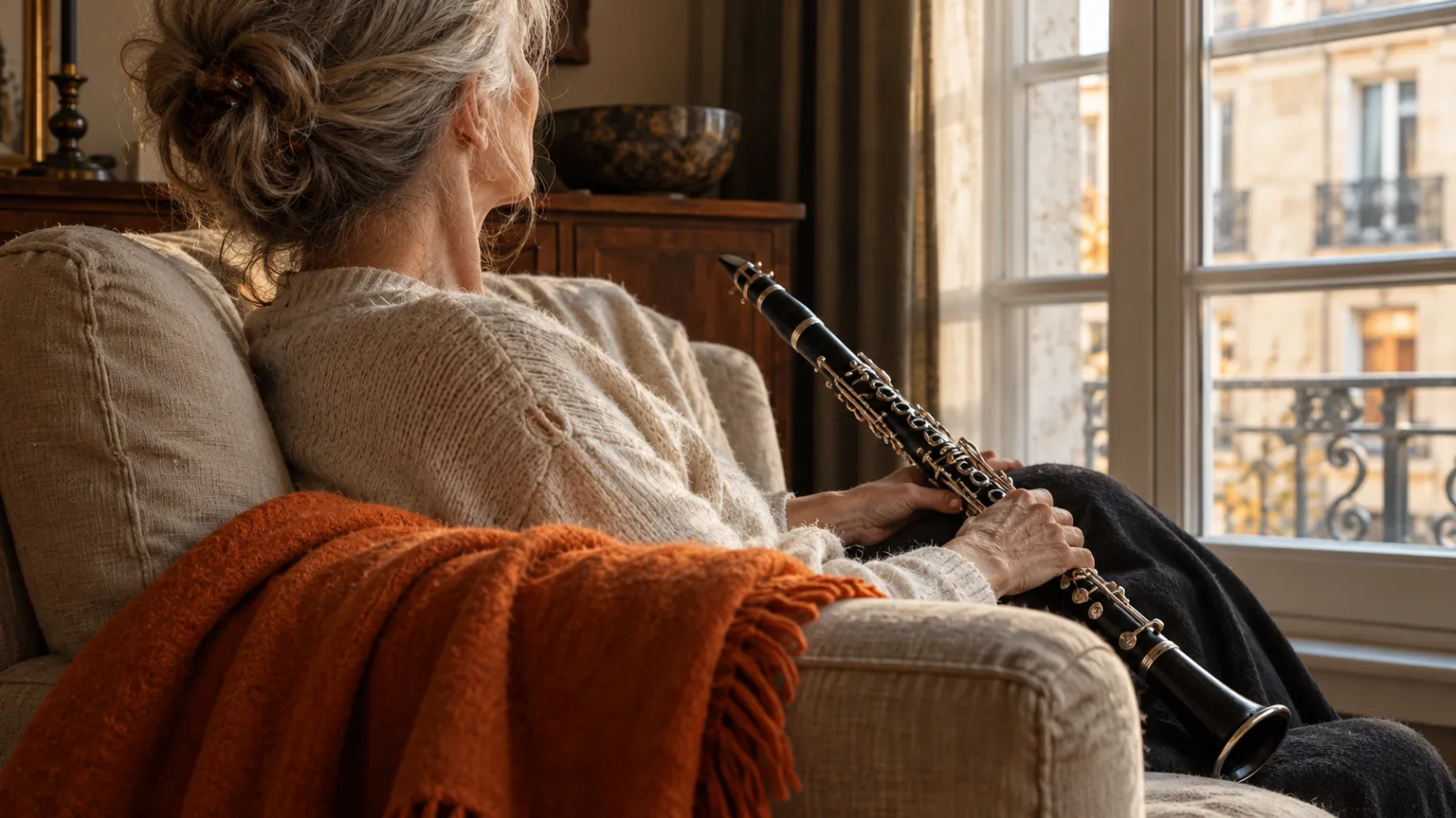 A woman in her early sixties sits in a linen armchair by a tall window, seen from behind, a wooden clarinet resting across her lap, with an orange wool throw over the chair arm in soft morning light.